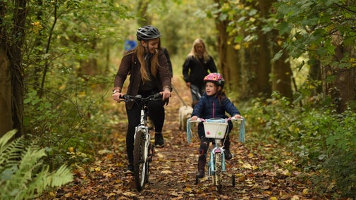 An adult and child cycling along the Tramway Trail surrounded by trees, with autumn leaves on the ground and dog walkers in the background.